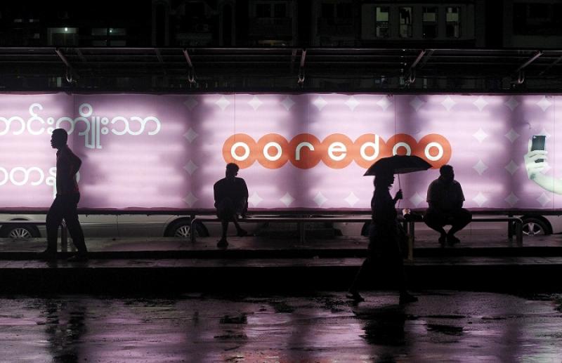 People sit at a bus station with an Ooredoo advertisement as they wait for a bus in Yangon August 1, 2014. REUTERS/FILE PHOTO
