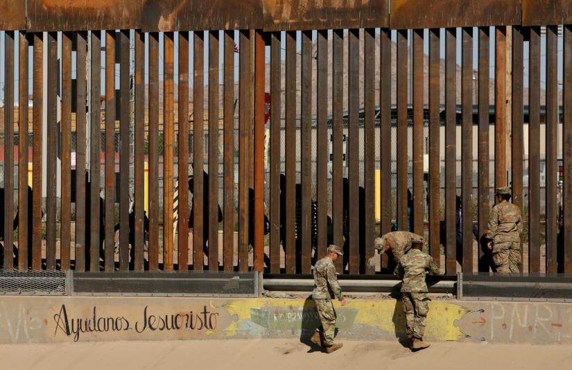 U.S. soldiers walk next to the border fence between Mexico and the United States, as migrants are seen walking behind the fence, after crossing illegally into the U.S. to turn themselves in, in El Paso, Texas, U.S., in this picture taken from Ciudad Juarez, Mexico, April 3, 2019. The writing on the wall reads, `Help us Jesus Christ.` REUTERS.