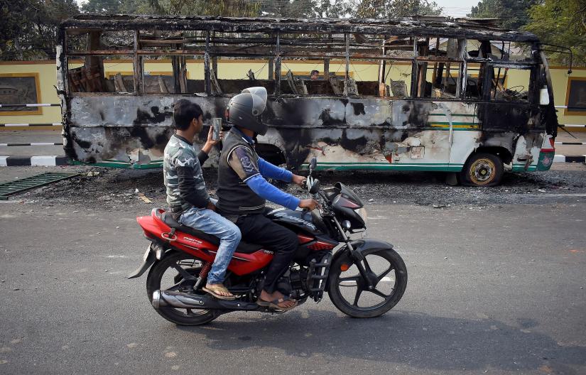 Men ride a motorcycle past a damaged bus that was set on fire by demonstrators, during a protest after India`s parliament passed Citizenship Amendment Bill, in Guwahati, India December 12, 2019. REUTERS